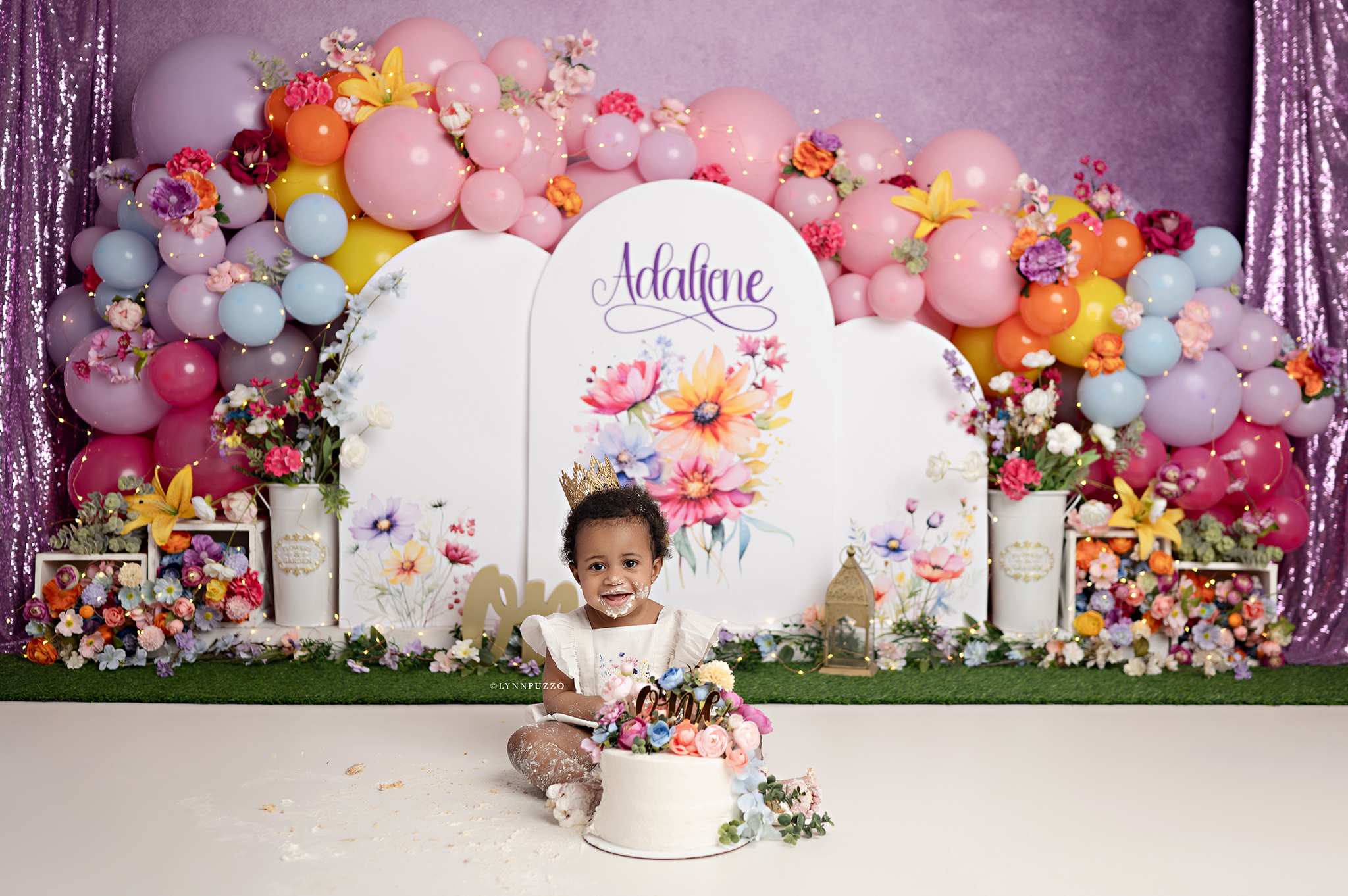 Wildflower cake smash featuring a baby girl in a white floral dress with a gold crown surrounded by a bright, colorful floral set